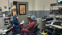 Physician educator converses with two trainees. All are masked and wearing scrubs.